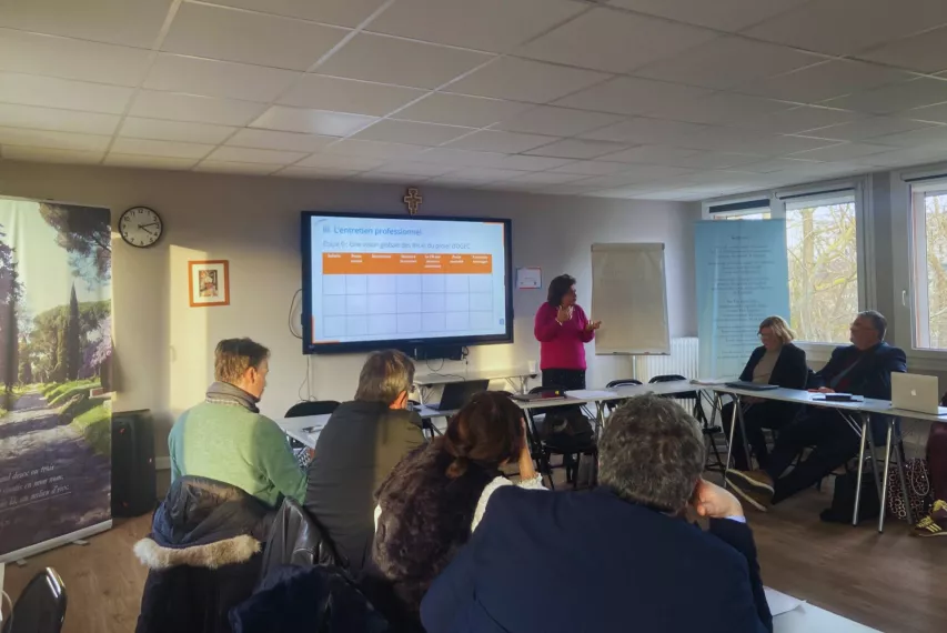 Des personnes sont assises autour d'une table et écoutent une personne debout devant un écran. 